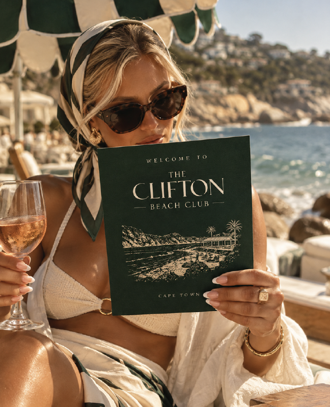 A member in a silk headscarf reading her Clifton Beach Club welcome card over a glass of rosé